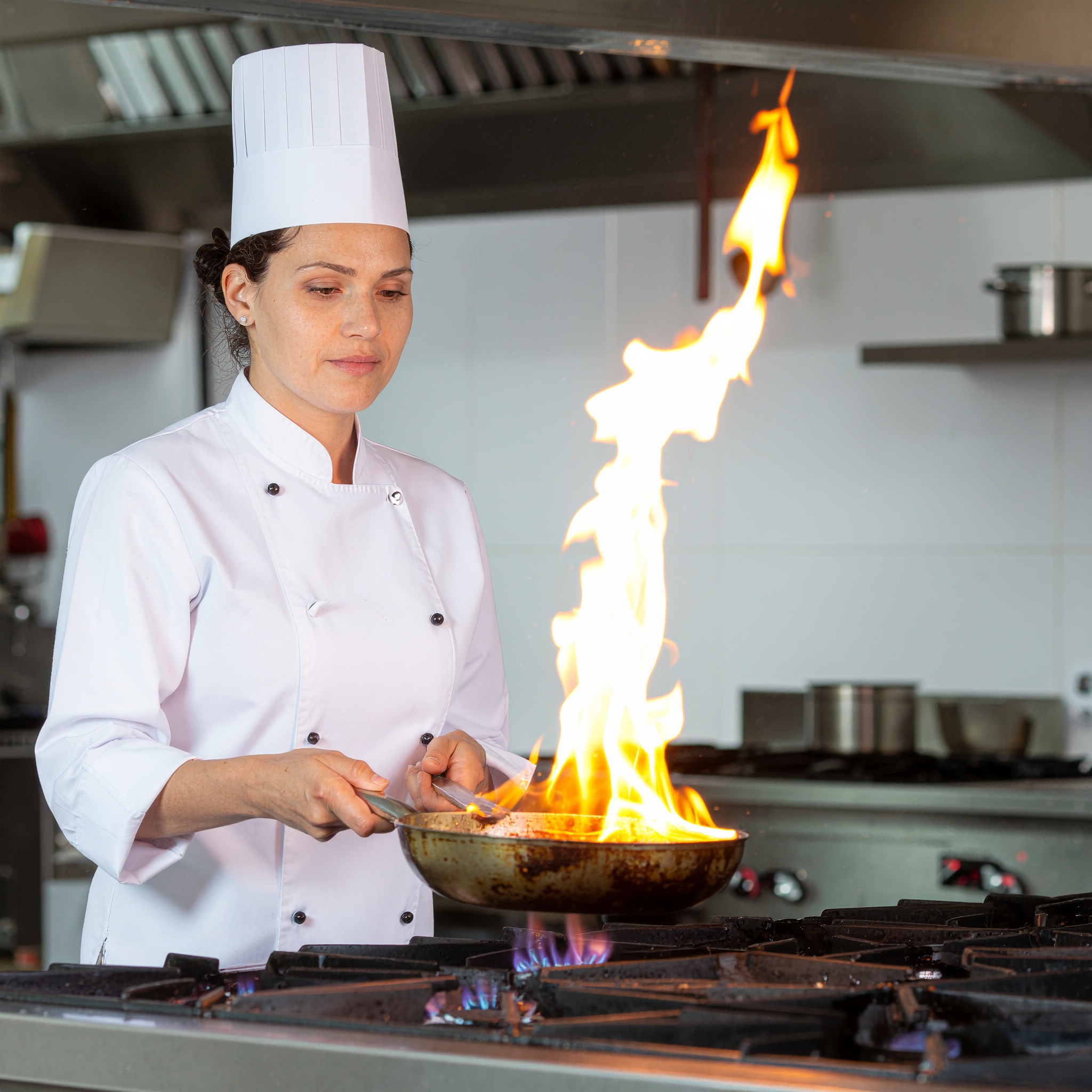 Chef de uniforme branco com chapéu alto segurando frigideira em chamas, em cozinha profissional com fundo metálico e iluminação brilhante.